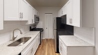 This single-floor residence is designed with a clean, neutral palette of whites and soft grays, creating a bright and cohesive atmosphere throughout. The layout flows logically from the open living area into the adjoining spaces, with plush wall-to-wall carpeting covering the main living and bedroom areas, while warm hardwood-style flooring runs through the kitchen and bathroom.

The kitchen is a standout feature, appointed with crisp white shaker cabinetry, granite-style countertops, a subway tile backsplash, and a full suite of stainless steel appliances including a gas range, microwave, and refrigerator. A galley-style configuration maximizes efficiency while maintaining a polished, modern look.

The living area is generously proportioned and benefits from abundant natural light through large sliding glass doors fitted with vertical blinds, offering views to the outdoors. Additional rooms include well-sized bedrooms, each featuring plush carpet and louvered closet doors providing ample storage space.

The bathroom is finished in a clean, contemporary style with a white vanity, undermount sink, a tub-shower combination, and hardwood-style flooring, completing the space with a tidy, functional design.