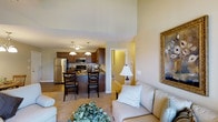 This single-floor space showcases an open-concept layout that seamlessly connects the living area and kitchen, creating a cohesive and functional environment. The living area is warm and inviting, furnished with light-toned seating accented by decorative pillows, and features notably high ceilings that lend an airy, spacious feel. A framed artwork on the wall and a classic table lamp add character and warmth to the space.

The kitchen is well-appointed, featuring rich dark wood cabinetry paired with dark granite countertops and stainless steel appliances, including a full-size refrigerator and built-in microwave. A peninsula with bar-height seating accommodates two dark wood barstools, making it an ideal spot for casual dining or entertaining.

Pendant lighting hangs above the kitchen peninsula, providing focused task lighting while complementing the overall warm ambiance of the space. Neutral-toned walls and carpeting throughout help unify the open layout, while a doorway visible in the background hints at additional rooms beyond. The overall atmosphere is comfortable and well-designed, balancing practicality with a polished aesthetic.