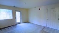 This single-floor space presents a clean, open layout with a straightforward and functional design. The main room features wall-to-wall carpeting in a neutral tone, transitioning to a hard surface flooring near the exterior door, providing a practical entryway area. The walls are painted in a soft, warm off-white, contributing to a bright and airy atmosphere. A glass-paneled exterior door with a built-in blind allows natural light to filter into the room, enhancing the overall sense of openness. The standard-height ceiling is smooth and white, fitted with a ceiling-mounted smoke detector or sprinkler fixture. Electrical outlets are positioned along the walls, and a wall switch plate is visible on one side, indicating readiness for customization and furnishing. A red fire alarm or alert device is mounted on the upper wall, reflecting attention to safety standards. The room is currently vacant, offering a versatile blank canvas that can be adapted to a variety of uses. Overall, the space is simple, well-lit, and move-in ready.