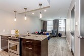 This single-floor unit showcases a thoughtfully designed open-concept layout that seamlessly connects the living area to the kitchen. The living space features light wood-look flooring, recessed lighting, and a large window that allows natural light to pour in while offering exterior views, creating a bright and welcoming atmosphere.

The kitchen is a standout feature, boasting dark espresso cabinetry paired with granite countertops, a tile backsplash, and stainless steel appliances including a refrigerator and microwave. A substantial island with bar seating serves as both a prep space and casual dining area, illuminated by modern pendant lights overhead.

The bedroom is a serene retreat, featuring a ceiling fan, hardwood-style flooring, and a generous window with scenic views. A sleek sliding barn-style door leads to a well-organized closet, while a gallery wall of framed black-and-white photography adds a sophisticated decorative touch.

The bathroom continues the refined aesthetic with dark cabinetry, granite countertops, a wide mirrored vanity, and clean modern fixtures. The space is finished with recessed lighting and an open layout that enhances its functional appeal. Throughout, neutral tones and contemporary finishes create a cohesive, polished living environment.