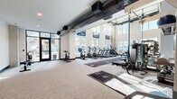 Located at 4312 Yancey Rd in Charlotte, NC, this fitness center offers a generously sized, single-floor exercise space designed to accommodate a wide variety of workout needs. The facility immediately impresses with its soaring double-height ceilings, exposed industrial-style ductwork, and expansive floor-to-ceiling windows that flood the interior with natural light and offer views of the surrounding community.

The main workout area features a long turf sprint/sled track marked with yardage lines, providing a dedicated space for functional and high-intensity training. A full row of treadmills, ellipticals, and stair climbers lines one side of the room, all equipped with personal screens. Strength training is well-supported with a cable machine, a power rack, a dumbbell rack, and mirrored walls that enhance the sense of space.

A dedicated cardio alcove near the glass entry doors houses rowing machines and stationary cycling bikes, bathed in natural light. A separate training zone toward the rear of the facility offers a suspension training rig and a full set of kettlebells and medicine balls, alongside organized storage shelving. A mezzanine level overlooks the main floor, adding architectural depth. The light, neutral color palette and carpet flooring create a clean, professional, and energizing workout environment throughout.
