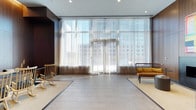 This single-floor space makes a striking first impression with its expansive floor-to-ceiling windows that flood the interior with natural light. Sheer, layered curtains soften the incoming sunlight while framing sweeping views of the surrounding urban skyline, creating an airy and refined atmosphere throughout.

The flooring features large-format tiles in a neutral, light tone, complemented by area rugs that define distinct zones within the open layout. The carefully considered design balances hard and soft surfaces to create a welcoming yet polished environment.

Furnishings are minimal and thoughtfully curated, including a classic wooden rocking chair and a sleek, contemporary bench with a warm mustard-toned cushion. These pieces reflect a blend of timeless and modern design sensibilities, contributing to the space's calm and inviting character.

The ceiling is clean and unadorned, with recessed lighting providing supplemental illumination alongside the generous natural light. Dark wall paneling on one side provides a bold contrast to the brightness of the windowed facade, adding depth and visual interest to the overall composition.

The result is a sophisticated, open interior that feels both functional and refined, ideal for a reception area, lounge, or collaborative gathering space in a contemporary commercial or mixed-use building.