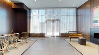 This single-floor space makes a striking first impression with its expansive floor-to-ceiling windows that flood the interior with natural light. Sheer, layered curtains soften the incoming sunlight while framing sweeping views of the surrounding urban skyline, creating an airy and refined atmosphere throughout.

The flooring features large-format tiles in a neutral, light tone, complemented by area rugs that define distinct zones within the open layout. The carefully considered design balances hard and soft surfaces to create a welcoming yet polished environment.

Furnishings are minimal and thoughtfully curated, including a classic wooden rocking chair and a sleek, contemporary bench with a warm mustard-toned cushion. These pieces reflect a blend of timeless and modern design sensibilities, contributing to the space's calm and inviting character.

The ceiling is clean and unadorned, with recessed lighting providing supplemental illumination alongside the generous natural light. Dark wall paneling on one side provides a bold contrast to the brightness of the windowed facade, adding depth and visual interest to the overall composition.

The result is a sophisticated, open interior that feels both functional and refined, ideal for a reception area, lounge, or collaborative gathering space in a contemporary commercial or mixed-use building.
