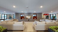 This expansive amenity space is thoughtfully designed to offer a wide range of recreational and social experiences across a single level. The centerpiece of the main lounge area is a sleek gas fireplace set into a striking patterned accent wall featuring a bold geometric tile design in muted blues and grays. Flanking the fireplace are wood-framed display niches with flat-screen monitors, adding a modern entertainment element to the space. Generous cream-toned sectional sofas and lounge chairs are arranged to encourage gathering, all set atop light tile and warm hardwood flooring.

Beyond the lounge, the space opens into a recreation area equipped with a bowling lane branded with vibrant overhead displays, as well as a ping-pong table, creating a lively, social atmosphere. Built-in shelving units along one wall provide stylish storage and display space. A kitchenette features gray shaker cabinetry, white countertops, stainless steel appliances including a full refrigerator and microwave, offering functional convenience.

Large floor-to-ceiling windows and glass doors throughout the space flood the interior with natural light and provide views of the surrounding outdoor areas, enhancing the open, inviting ambiance of this well-appointed communal space.