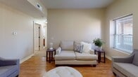 This single-floor residence offers a thoughtfully arranged layout that balances comfort and functionality across several distinct spaces. The living area features warm hardwood-style flooring, neutral beige walls, and generous ceiling height, creating an open and inviting atmosphere with easy flow to the rest of the home. A dedicated den or office space is carpeted and benefits from natural light streaming through multiple windows fitted with blinds, making it an ideal area for remote work or study. The bedroom is carpeted and features dual windows that allow for ample natural light, lending a calm and restful ambiance. The kitchen is efficiently designed with rich wood cabinetry, granite-style countertops, stainless steel appliances including a full-size refrigerator, an electric range, and a built-in microwave, along with a distinctive glass block window that adds a unique architectural detail. The bathroom is well-finished with tile flooring, a full soaking tub and shower combination, a wood-framed medicine cabinet, and a vanity with granite-style countertops. Throughout the residence, consistent warm beige tones and clean finishes create a cohesive and welcoming environment suited to a variety of lifestyles.