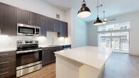 This contemporary single-floor space showcases a thoughtfully designed open-concept layout with clean lines and modern finishes throughout. The kitchen serves as the centerpiece, featuring rich dark-toned shaker-style cabinetry that extends to the ceiling, white quartz countertops, and a full suite of stainless steel appliances including a refrigerator, range, microwave, and dishwasher. A white kitchen island with pendant lighting provides additional prep space and a natural gathering point, while a classic white subway tile backsplash and under-cabinet lighting complete the polished look. Light hardwood flooring flows seamlessly throughout, lending warmth and continuity to the open floor plan. The living area is bright and airy, benefiting from soaring ceilings, a ceiling fan, and expansive floor-to-ceiling windows with sliding balcony access that flood the space with natural light and offer views of the surrounding streetscape. The bathroom mirrors the kitchen's design aesthetic, with matching dark cabinetry, white quartz countertops, a framed mirror, and contemporary vanity lighting. Tall storage cabinetry in the bathroom provides ample organization space. The overall ambiance is clean, modern, and move-in ready, with a cohesive design palette that balances style and functionality.