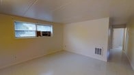 This single-floor space offers a straightforward and functional layout designed for versatility. The interior is characterized by light cream-colored walls that contribute to a bright and neutral ambiance throughout. The flooring consists of light-toned tile, which is easy to maintain and adds a clean, uniform appearance to the space. The ceiling is finished with a flat, paneled surface that keeps the room feeling tidy and well-contained. A window fitted with horizontal blinds allows for natural light to filter in while providing privacy control. Beneath the window, a window-mounted air conditioning unit ensures comfortable climate regulation during warmer months. Complementing this, a wall-mounted floor vent provides additional climate control, suggesting a heating system is also in place. Electrical outlets are positioned along the walls for convenient access. The open, unobstructed floor plan offers flexibility for a variety of uses, whether as a living area, workspace, or storage. Overall, the space presents a clean, no-frills environment that is practical and ready to be configured to suit a range of needs.