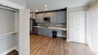 This single-floor studio offers a thoughtfully organized open-concept layout that maximizes every square foot. The main living area features rich, dark hardwood-style flooring, recessed lighting, and a ceiling fan, creating a comfortable and functional environment. Crown molding and neutral gray walls contribute to a clean, contemporary aesthetic throughout. Windows with vertical blinds allow natural light to fill the space.

The kitchen runs along one wall and is well-appointed with gray shaker-style cabinetry extending to the ceiling for ample storage, a textured tile backsplash, light countertops, and a full suite of stainless steel appliances including a refrigerator, microwave, dishwasher, and cooktop. A built-in desk area adjacent to the kitchen adds a practical workspace element.

The bathroom is finished with large-format beige tile flooring, gray cabinetry with a single vanity and mirror, and a toilet — offering a clean, modern feel consistent with the rest of the unit. The overall design maintains a cohesive neutral palette of grays and warm wood tones, delivering a polished and functional living space.