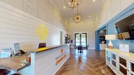 The Grand Life leasing office is a thoughtfully designed, single-floor space that blends modern aesthetics with warm, welcoming details. Upon entry, visitors are greeted by a prominent reception desk adorned with the Grand Life logo in glowing neon, set against shiplap-clad walls that extend dramatically upward toward soaring ceilings. Striking sputnik-style chandeliers cast warm light across the herringbone-patterned hardwood floors, creating an inviting and upscale atmosphere throughout.

The open layout flows naturally from the reception area into a casual seating lounge, where a round table and leather chairs offer a relaxed setting for conversation. A blue-accented accent wall with diagonal shiplap detailing frames a cozy nook housing a coffee and refreshment station, adding a pop of color to the otherwise neutral palette. Decorative shelving, tasteful accessories, and branded touches are thoughtfully placed throughout.

A dedicated meeting room, enclosed with sleek black-framed glass doors, provides a professional setting equipped with a conference table and mounted display. Adjacent to this is a smaller seating area bathed in natural light from floor-to-ceiling curtained windows, offering an airy and comfortable spot for consultations. Together, the spaces combine functionality with refined design to create a polished and hospitable environment.