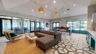 This single-floor amenity space blends contemporary design with functional versatility, offering a well-appointed environment suited for both productivity and leisure. The main lounge area is anchored by a tufted sectional sofa set atop a bold geometric-patterned tile floor, complemented by woven rattan pendant lights that hang from elevated ceilings, adding warmth and character to the space. A striking deep blue accent wall with a wall-mounted flat-screen TV serves as a focal point in the lounge, framed by botanical artwork and decorative accessories that reinforce a nature-inspired aesthetic. Adjacent to the lounge, a kitchenette area features vibrant teal cabinetry, open wooden shelving set against a navy diamond-tile backsplash, stainless steel appliances, and a dark high-top bar with red bar stools — all bathed in natural light streaming through teal-framed windows. Multiple glass-walled conference rooms, numbered and clearly designated, provide private meeting spaces with ample seating and direct views to the outdoors. Light wood-tone flooring flows through the transitional corridors, while decorative wall art adds artistic interest throughout. The space is bright, cohesive, and thoughtfully designed to serve as a dynamic hub for community members.
