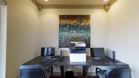 This single-floor business center is a functional and thoughtfully appointed workspace designed to meet professional needs. The room is anchored by a dark, wide-surface desk that comfortably accommodates two computer workstations, each equipped with a monitor and keyboard, along with a centrally positioned Xerox multifunction printer for scanning, copying, and printing needs. Ergonomic mesh chairs are provided at each station, ensuring comfort during use. Recessed ceiling lighting delivers consistent, even illumination throughout the space, while decorative crown molding along the ceiling adds a refined architectural touch. A large, vibrant abstract painting mounted on the central wall serves as a striking focal point, incorporating rich tones of teal, gold, and brown that bring warmth and visual interest to the otherwise neutral interior. The walls feature a soft, light finish that complements the overall clean and professional ambiance. This space is well-suited for guests or tenants requiring access to business resources in a quiet, organized setting.