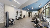 This fitness room at 1559 NW 15th Street Rd in Miami, FL offers a thoughtfully designed exercise space that combines modern aesthetics with comprehensive workout functionality. The room features a striking two-tone ceiling design, with a bold steel-blue accent contrasting against crisp white sections, illuminated by sleek linear LED lighting that ensures bright, even coverage throughout. A decorative marble-patterned feature wall adds a refined touch to the otherwise clean, white interior. Large windows along one side of the room invite abundant natural light and provide views of the surrounding greenery, creating an energizing atmosphere. The durable speckled rubber flooring provides both safety and comfort underfoot. Equipment is well-organized and spans both cardio and strength training categories. The cardio section includes multiple treadmills, an elliptical trainer, and a rowing machine, all positioned to take advantage of the natural light and outdoor views. The strength training area is anchored by a multi-functional cable machine and a power rack with weight plates, complemented by a tiered dumbbell rack offering a range of free weights. Full-length mirrors on one wall enhance the sense of space and allow users to monitor their form. The room is well-ventilated with visible ceiling vents, and the open layout ensures comfortable circulation throughout the space.