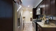 This single-floor space showcases a thoughtfully designed interior centered around a striking galley-style kitchen. The kitchen is outfitted with rich, dark espresso-toned wood cabinetry that extends from floor to ceiling, providing generous storage while creating a bold visual contrast against the light cream-colored walls and countertops. Stainless steel appliances — including a French door refrigerator with an in-door dispenser, a gas range, and an over-the-range microwave — lend a polished, contemporary feel to the space. A decorative tile backsplash in cool blue-gray tones adds a subtle layer of texture and color behind the range. The light wood-look flooring runs the length of the kitchen, contributing warmth and continuity underfoot. Recessed ceiling lighting provides clean, even illumination throughout. The galley layout is efficient and well-organized, with countertop space on both sides creating an ideal workflow for cooking and meal preparation. At the far end of the kitchen, a glimpse into an adjacent room hints at additional living space beyond, suggesting a well-connected and cohesive floor plan. Overall, the space balances functionality with a modern aesthetic through its carefully selected finishes and fixtures.