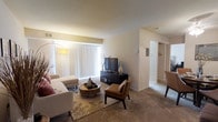 This single-floor residence offers a well-organized and comfortable layout designed for everyday living. The living room serves as the central gathering space, featuring plush wall-to-wall carpeting that adds warmth underfoot. A large sliding glass door fitted with vertical blinds allows an abundance of natural light to fill the room, creating a bright and welcoming atmosphere. The space accommodates a generous seating arrangement, complemented by a media console and tasteful décor that enhance the room's contemporary feel. A tall arc floor lamp adds a stylish ambient lighting option. The neutral, warm-toned walls provide a versatile backdrop throughout the unit. A hallway visible from the living area leads to additional rooms, suggesting a practical and connected floor plan. The transition between spaces is smooth and open, contributing to an overall sense of spaciousness. The layout appears to include access to a closet or storage area, adding to the unit's functionality. Overall, the space presents a clean, move-in-ready interior with a modest yet modern aesthetic suited for comfortable residential living.
