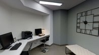 This single-floor office space offers a streamlined, functional environment designed for productivity. The room is finished in neutral grey tones throughout the walls and carpet, creating a calm and professional atmosphere. A built-in white floating desk runs along one wall, seamlessly integrated into a recessed nook that provides a defined workspace. The desk accommodates a desktop computer, printer, and desk lamp, with sleek black task chairs on chrome bases completing the workstation setup. Overhead, a recessed rectangular panel light provides bright, even illumination across the workspace. The ceiling features subtle architectural detailing with a stepped soffit above the desk area, adding a modest design element to the otherwise minimalist interior. A tufted white bench with a dark metal frame is positioned along the opposite wall, offering additional seating or a waiting area. Wall-mounted artwork provides a subtle decorative accent. The overall ambiance is clean, modern, and understated, with a focus on practicality and efficient use of space.