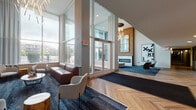 Located at 212 W Saint Charles Rd in Lombard, Illinois, this contemporary lobby entrance makes a striking first impression with its dramatic double-height ceilings and expansive floor-to-ceiling glass windows that flood the space with abundant natural light and offer open views of the surrounding streetscape. The flooring throughout features a distinctive herringbone-patterned hardwood that extends seamlessly from the entrance vestibule into the main corridor, lending warmth and visual continuity to the space.

The lobby is organized into several well-defined seating areas, each thoughtfully furnished with modern lounge chairs, leather sofas, and accent tables. A standout fireplace set against a natural wood-clad accent wall serves as a focal point, complemented by a large mirror and bold contemporary artwork. Sculptural, branch-like chandeliers add an artistic lighting element overhead.

Additional amenities visible within the lobby include a resident mailbox area with individual secure units and a wall-mounted flat-screen display near the reception desk area. A long central corridor with herringbone flooring leads residents deeper into the building, lined with recessed lighting for a clean, polished aesthetic. The overall ambiance balances modern design with warm, natural materials to create an inviting and functional communal environment.