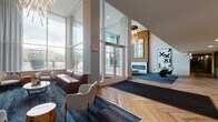 Located at 212 W Saint Charles Rd in Lombard, Illinois, this contemporary lobby entrance makes a striking first impression with its dramatic double-height ceilings and expansive floor-to-ceiling glass windows that flood the space with abundant natural light and offer open views of the surrounding streetscape. The flooring throughout features a distinctive herringbone-patterned hardwood that extends seamlessly from the entrance vestibule into the main corridor, lending warmth and visual continuity to the space.

The lobby is organized into several well-defined seating areas, each thoughtfully furnished with modern lounge chairs, leather sofas, and accent tables. A standout fireplace set against a natural wood-clad accent wall serves as a focal point, complemented by a large mirror and bold contemporary artwork. Sculptural, branch-like chandeliers add an artistic lighting element overhead.

Additional amenities visible within the lobby include a resident mailbox area with individual secure units and a wall-mounted flat-screen display near the reception desk area. A long central corridor with herringbone flooring leads residents deeper into the building, lined with recessed lighting for a clean, polished aesthetic. The overall ambiance balances modern design with warm, natural materials to create an inviting and functional communal environment.
