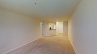 This single-floor space offers a clean, open layout designed for comfortable everyday living. The main living area is finished with plush wall-to-wall carpeting in a neutral tone, creating a warm and inviting atmosphere throughout. Smooth, light-colored walls and white trim contribute to a bright, airy feel, while recessed lighting provides consistent illumination across the space.

A partial wall with a pass-through counter separates the living area from the kitchen, allowing for an open, connected feel between the two spaces. The kitchen area is visible through the pass-through, where a mirror or window reflects additional light, enhancing the sense of openness. White cabinetry and neutral countertops complete the kitchen's clean aesthetic.

A hallway extends toward the rear of the unit, lined with white doors that suggest access to additional rooms such as a bathroom or storage areas. The corridor is well-lit, maintaining the consistent brightness found throughout the space. An entry door is visible at the end of the hallway, reinforcing the practical flow of the layout.

Overall, the space presents a straightforward and functional design with neutral finishes, making it highly adaptable to a variety of interior styles and furnishing choices.