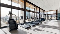 This expansive fitness center offers a premium exercise experience within a thoughtfully designed, single-floor layout. The space is defined by dramatic floor-to-ceiling glass curtain walls that span nearly the entire perimeter, flooding the interior with abundant natural light and framing sweeping panoramic views of the surrounding urban skyline. Warm wood-paneled ceiling accents complement the crisp white tile grid ceiling, creating a refined yet energizing atmosphere.

The exercise room is comprehensively equipped to accommodate a wide range of fitness routines. A long row of treadmills and elliptical trainers lines the windowed walls, allowing users to exercise while taking in the city views. Dedicated zones house a full array of Hoist and other commercial-grade strength machines, a well-stocked dumbbell rack ranging across multiple weight increments, and a free-weight area complete with power racks, barbells, and adjustable benches. Large mirrors along interior walls enhance the sense of space and support form monitoring during workouts.

Built-in open shelving provides convenient storage, while a wall-mounted flat-screen television adds an entertainment element. The flooring transitions from large-format tile in the cardio zones to rubber sport flooring in the strength areas, ensuring both safety and functionality. Access to an adjacent outdoor terrace, furnished with seating, further elevates the overall experience of this high-end fitness facility.