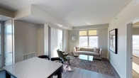 This single-floor residence showcases a well-designed open-concept layout that seamlessly connects the kitchen and living areas. The living room is bathed in natural light courtesy of multiple large windows, including a sliding glass door that opens to an outdoor balcony, offering views of the surrounding landscape. Neutral gray walls and smooth ceilings create a clean, modern backdrop throughout the space. Dark hardwood-style flooring runs throughout, adding warmth and contrast to the light color palette. The living area is anchored by a plush area rug, a glass coffee table, and a beige sofa accented with decorative pillows, complemented by a green accent chair and round side table. A decorative sunburst mirror serves as a focal point on the wall, adding a touch of elegance. The kitchen area, visible in the foreground, features a granite-style countertop with bar seating, providing a functional and stylish transition between spaces. Overall, the unit presents a bright, contemporary ambiance with thoughtful design details and a cohesive neutral tone throughout.