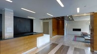 Located at 10 DeKalb Avenue in White Plains, New York, this single-floor commercial space presents a polished and contemporary aesthetic designed to make a strong first impression. The reception area serves as the centerpiece of the space, featuring a striking front desk with warm wood cabinetry and a clean white illuminated panel, creating a welcoming yet professional focal point.

The interior design balances contrasting materials with great effect — rich wood wall paneling frames a prominent entryway, while a bold dark tile accent wall adds depth and visual interest. The flooring features a distinctive mix of light and darker wood-tone planks arranged in a geometric pattern, adding a dynamic element to the overall design.

Recessed and linear LED lighting is embedded throughout the ceiling, providing bright, even illumination across the space while enhancing its modern character. A fireplace is visible in the background, lending a refined warmth to the otherwise crisp, minimalist environment. The open layout and thoughtful material palette create a sophisticated ambiance that is well-suited for a professional office or upscale business setting.