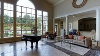 This sophisticated single-floor space makes a striking first impression with its soaring double-height ceilings and dramatic floor-to-ceiling arched windows that flood the interior with natural light. The expansive windows frame views of a sparkling outdoor pool surrounded by mature trees, creating a seamless connection between interior and exterior. Decorative white columns with classical detailing define the space and complement the refined architectural character throughout. The flooring features an elegant combination of light tile and rich dark marble inlay, adding visual interest and a sense of grandeur underfoot. A grand black baby grand piano anchors one side of the room, lending a refined, upscale ambiance. Nearby, a small dining arrangement with dark wood chairs provides an intimate seating option. On the opposite side, a tufted camel-toned sofa with coordinating accent pillows is paired with glass-topped coffee tables, creating a comfortable lounge area. Crown molding, recessed lighting, and neutral warm-toned walls complete the polished aesthetic. Overall, the space exudes a formal yet welcoming atmosphere, ideal for elegant gatherings or leisurely relaxation.