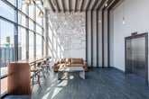 This contemporary lobby space makes a striking first impression with its dramatic floor-to-ceiling glass curtain wall, which floods the interior with abundant natural light and offers unobstructed views of the surrounding streetscape. The soaring ceiling is defined by a bold arrangement of parallel dark wood beams that run horizontally across the space, adding warmth and architectural character while complementing the dark-toned wood-look tile flooring below.

A standout design element is the large-scale city map mural adorning one wall, referencing the local area and serving as a focal point that grounds the space in its community context. Vertical dark wood slat paneling flanks the mural wall, reinforcing the space's cohesive industrial-modern aesthetic.

The lobby is thoughtfully appointed with multiple seating areas. A cognac leather sofa paired with a minimalist concrete coffee table offers a relaxed lounge setting, while a high-top table with industrial-style adjustable bar stools provides a more functional workspace near the windows. A cozy nook features a pair of plush blue velvet swivel chairs flanking a small accent table, adding a vibrant pop of color.

Edison-style pendant bulbs suspended from the ceiling contribute an ambient, inviting warmth. A stainless steel elevator provides convenient vertical access throughout the building, completing this well-designed and welcoming lobby.