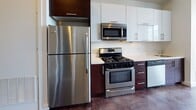 Located at 66–70 Beale St in Quincy, Massachusetts, this single-floor unit presents a clean, contemporary design characterized by polished concrete floors that extend consistently throughout the space, lending an industrial-modern aesthetic. The open living area is anchored by large, black-framed windows that invite abundant natural light and offer views of the surrounding streetscape, creating a bright and airy atmosphere.

The kitchen is well-appointed with a full suite of stainless steel appliances, including a gas range, over-the-range microwave, refrigerator, and dishwasher. Two-toned cabinetry—white upper cabinets paired with dark lower cabinets—provides a striking visual contrast against the light countertops and white tile backsplash, offering ample storage and a functional workspace.

A separate bedroom area features a distinctive interior clerestory window, allowing light to pass between spaces while maintaining definition between rooms. The bathroom includes a bathtub with shower, toilet, and grab bar, finished with the same polished concrete flooring found throughout the unit. White walls are used consistently across all areas, contributing to a cohesive, minimalist character throughout the space.