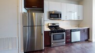 Located at 66–70 Beale St in Quincy, Massachusetts, this single-floor unit presents a clean, contemporary design characterized by polished concrete floors that extend consistently throughout the space, lending an industrial-modern aesthetic. The open living area is anchored by large, black-framed windows that invite abundant natural light and offer views of the surrounding streetscape, creating a bright and airy atmosphere.

The kitchen is well-appointed with a full suite of stainless steel appliances, including a gas range, over-the-range microwave, refrigerator, and dishwasher. Two-toned cabinetry—white upper cabinets paired with dark lower cabinets—provides a striking visual contrast against the light countertops and white tile backsplash, offering ample storage and a functional workspace.

A separate bedroom area features a distinctive interior clerestory window, allowing light to pass between spaces while maintaining definition between rooms. The bathroom includes a bathtub with shower, toilet, and grab bar, finished with the same polished concrete flooring found throughout the unit. White walls are used consistently across all areas, contributing to a cohesive, minimalist character throughout the space.