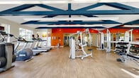 Located at 1456 Serfas Club Dr in Corona, California, this single-story fitness and recreation facility offers a comprehensive range of amenities designed to support an active lifestyle. The main workout area is a standout feature, boasting soaring vaulted ceilings accented by bold navy blue exposed beam trusses that lend the space an open, energetic atmosphere. Wood-look flooring runs throughout, complementing the vibrant orange accent walls that add a sense of motivation and energy. The gym floor is well-equipped with treadmills, strength training machines, free weights, and cable equipment, all reflected in full-length mirrors that enhance the sense of spaciousness. Glass partition walls and sliding glass doors separate distinct workout zones, allowing natural light to flow freely while maintaining visual connectivity across the space. An adjacent indoor court serves dual purposes, accommodating both basketball and racquetball, with a polished hardwood floor, painted court lines, and a wall-mounted basketball hoop. The facility also includes well-appointed locker rooms featuring richly textured multi-toned slate tile on floors, walls, and open shower areas, providing a durable and refined finish. Recessed lighting throughout ensures consistent illumination across all areas of this well-designed recreational facility.