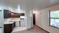 This single-floor residential unit offers a practical and straightforward layout designed for comfortable everyday living. The kitchen is a standout functional space, outfitted with rich dark wood cabinetry that provides ample storage, white appliances including a range, dishwasher, and full-size refrigerator, and light-toned tile flooring that adds brightness to the area. A double sink is positioned beneath overhead cabinets, maximizing workspace efficiency. The kitchen flows naturally into an adjacent carpeted area, which transitions toward the main entry door — a classic dark wood panel door that anchors the space with a welcoming presence. The neutral wall tones throughout create a clean, understated backdrop that complements the darker cabinetry accents. Flush-mount ceiling lighting provides consistent illumination across the kitchen and adjoining spaces. The open sightlines between the kitchen and the entry area contribute to a sense of connected, accessible space, making the layout easy to navigate. The combination of tile in the kitchen and carpet in the living areas provides distinct functional zones while maintaining a cohesive overall aesthetic throughout this well-organized single-level unit.
