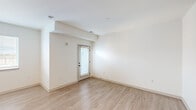 This single-floor unit presents a clean, contemporary design defined by a neutral white palette, light wood-tone luxury vinyl plank flooring, and recessed lighting throughout. The open-concept kitchen serves as a functional centerpiece, outfitted with stainless steel appliances including a range, microwave, dishwasher, and refrigerator. Light wood cabinetry with matte black hardware pairs with a white tile backsplash and light countertops, creating a cohesive, modern aesthetic. An integrated built-in desk or counter extension provides additional workspace or dining utility directly adjacent to the kitchen. The living area is generously proportioned and bathed in natural light through a window and a glass-paneled exterior door fitted with blinds, offering both brightness and privacy. Structural ceiling beams add subtle architectural interest to the open living space. The bathroom maintains the same modern sensibility, featuring a single-sink vanity with wood cabinetry and black hardware, a tub-shower combination with brushed nickel fixtures, and wood-look tile flooring. A stacked laundry unit is also present within the unit. The overall ambiance is light, airy, and move-in ready, with consistent finishes creating a cohesive look throughout.