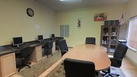 This single-floor office space is designed to support both individual productivity and group collaboration. The room features a built-in dark granite-topped workstation along one wall, equipped with two computer setups and accompanied by ergonomic task chairs, making it well-suited for focused desk work or administrative tasks. A utility cabinet is mounted to the wall nearby, providing convenient enclosed storage for office supplies or equipment.

At the center of the room sits a spacious oval conference table surrounded by multiple rolling office chairs, offering a dedicated area for meetings or teamwork. A freestanding bookcase along one wall adds additional open storage and display space.

The walls are finished in a soft, neutral yellow tone, lending a warm and approachable atmosphere to the workspace. Decorative wall art adds a touch of personality without overwhelming the professional setting. The floor transitions between tile and a patterned area rug beneath the workstation zone, adding texture and visual interest. Overhead fluorescent lighting ensures the space is well-illuminated throughout. A wall clock keeps the room practical and organized. Overall, this office presents a clean, efficient, and welcoming environment suited for a small business or shared professional setting.