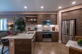 This thoughtfully designed single-floor residence seamlessly blends modern aesthetics with functional living. The open-concept kitchen serves as a focal point, showcasing rich wood-toned cabinetry paired with open floating shelves, a striking chevron-patterned white tile backsplash, and a suite of stainless steel appliances including a gas range, built-in microwave, and a French door refrigerator. A waterfall-edge kitchen island with bar seating provides an ideal space for casual dining and entertaining, all set atop warm light-wood flooring that flows throughout the main living areas.

The living room exudes a sophisticated, curated ambiance with warm taupe walls, recessed lighting, and a glass-paneled door that invites natural light and connects the interior to a private outdoor area. A large statement mirror amplifies the sense of space, while the overall design creates a welcoming, intimate atmosphere.

The bedroom offers a serene retreat, highlighted by soft neutral walls, plush carpeting, and windows that frame views of lush palm trees, allowing generous natural light to fill the room.

The bathroom is finished with large-format tile flooring, a floating wood vanity topped with a white countertop, a backlit mirror, and a glass-enclosed shower, delivering a clean, spa-inspired feel that complements the residence's modern character.