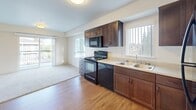 Located at 4112 133rd St SE in Mill Creek, Washington, this single-floor residence offers a thoughtfully designed layout that balances functionality and comfort. The kitchen serves as a central highlight, featuring rich dark wood cabinetry, light countertops, and a full suite of black appliances including an electric range, built-in microwave, dishwasher, and refrigerator. Hardwood-style flooring runs throughout the main living and kitchen areas, lending warmth and continuity to the space. Large windows allow generous natural light to pour in, offering pleasant views of the surrounding Pacific Northwest greenery. The living area transitions seamlessly from the kitchen, with plush carpeting underfoot and access to an outdoor balcony. The bedroom is carpeted and well-lit by a wide window framing lush outdoor views, creating a serene retreat. The bathroom is clean and efficiently arranged, with warm wood-toned cabinetry, a single sink vanity, hardwood-style flooring, and a full soaking tub with a bright white surround. The overall ambiance of the space is light, neutral, and move-in ready, with a cohesive design palette of warm tones and modern finishes throughout.