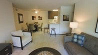 This single-floor residence offers a thoughtfully designed open-concept layout that seamlessly connects the living, dining, and kitchen areas into one cohesive and functional space. The living area is comfortably appointed with plush carpeted flooring in neutral tones, providing a warm and inviting atmosphere. A ceiling fan overhead ensures year-round comfort, while tasteful wall art and coordinated furnishings contribute to the space's polished aesthetic. Adjacent to the living area, the dining space accommodates a round table with upholstered chairs, making it ideal for both everyday meals and casual entertaining. The kitchen features a pass-through breakfast bar with bar stool seating, allowing for easy interaction between the kitchen and the main living areas. Dark wood cabinetry provides contrast against the lighter walls and countertops, and under-cabinet lighting adds a functional yet stylish touch. The open bar-style counter enhances the sense of connectivity throughout the space. Overall, the residence presents a clean, contemporary feel with a cohesive neutral color palette, warm lighting, and a layout that prioritizes both comfort and practicality for everyday living.