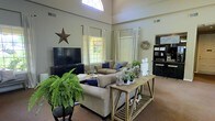 This single-floor space makes a striking impression with its generously proportioned living area, defined by dramatically high ceilings that lend an open, airy quality to the interior. A standout architectural feature is the arched transom window positioned near the top of the wall, allowing abundant natural light to pour into the room and draw the eye upward. Large paneled windows dressed with white curtains and blinds further brighten the space while offering views to the outdoors.

The living area is thoughtfully arranged with a substantial sectional sofa in a neutral taupe tone, accented with navy blue throw pillows that add a pop of color. A rustic, trestle-style console table serves as a decorative focal point behind the seating arrangement, styled with candles, greenery, and coastal-inspired accents. Lush Boston ferns and potted plants throughout the room bring a vibrant, natural element indoors.

A wall-mounted flat-screen television sits atop a dark media console, while a decorative metal star adorns the wall nearby, reinforcing a farmhouse-country aesthetic. A sunburst mirror and a built-in shelving or bar area are visible in the background, adding both function and visual interest. The warm-toned flooring and soft neutral wall color tie the entire space together, creating a welcoming and cohesive atmosphere.