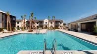 This amenity space combines an expansive outdoor pool area with a well-appointed fitness center, delivering a comprehensive recreational experience. The pool area features a generously sized swimming pool with clearly marked depth zones, surrounded by a clean, light-toned deck. Tall palm trees line the pool perimeter, evoking a resort-like atmosphere under open skies, while lounge seating and an outdoor dining area with a covered pavilion provide spaces to unwind. The backdrop of modern, white-painted multi-story buildings with dark-framed windows adds a polished architectural presence to the setting.

The fitness center is housed in a bright, single-floor interior with neutral walls, recessed lighting, and ceiling fans that maintain a comfortable environment. Herringbone-patterned carpet tiles cover the floor throughout. The space is divided into functional exercise zones: a cardio area features multiple treadmills positioned beneath wall-mounted TVs and large windows that allow generous natural light and outdoor views. The strength and functional training zone is equipped with a cable machine, power rack, rowing machines, Precor cycling equipment, a full dumbbell rack, medicine ball storage, battle ropes, and stability balls. Full-length mirrors along one wall visually expand the space and support proper form during workouts.