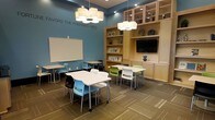 This single-floor space is designed as a collaborative study and learning environment, combining functional design with an energizing aesthetic. The room's most striking feature is its bold teal accent wall, which displays the motivational quote "Favors the Prepared Mind," setting an academic tone throughout. Pendant drum light fixtures hang from the ceiling, casting warm, even illumination across the space and complementing the recessed lighting above.

The layout accommodates multiple configurations, with white rectangular tables paired with a mix of colorful modern chairs in blue, green, and black, encouraging flexible seating arrangements for both individual study and group collaboration. A wall-mounted whiteboard provides a functional teaching or brainstorming surface.

Along one wall, a built-in shelving unit of light natural wood houses glass-front upper cabinets, open display shelves with decorative objects and greenery, and lower closed cabinetry. A flat-screen television is integrated into this built-in, adding a multimedia resource to the space. The patterned carpet tile flooring ties the room's neutral and vibrant tones together, contributing to a structured yet welcoming atmosphere.
