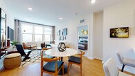 This well-appointed single-floor residence showcases a cohesive contemporary design with a neutral palette, light hardwood flooring, and an abundance of natural light throughout the main living areas. The open-concept layout seamlessly connects the dining area and living room, where a round marble-top dining table and mid-century modern chairs anchor one end, while a comfortable sectional sofa and curated furnishings define the lounge space. Large windows frame outdoor views, filling both areas with natural light.

The kitchen is outfitted with flat-front white lower cabinetry, warm wood upper cabinets with open shelving, white quartz countertops, and stainless steel appliances including a gas range and built-in microwave. Recessed lighting and a pendant light complete the functional yet stylish space.

The primary bedroom offers plush carpeting, generous window exposure, and decorative wall accents, accompanied by a spacious walk-in closet with wire shelving and carpet flooring. A second bedroom provides comfortable proportions with natural light and ample room for a workspace. The bathroom features large-format tile flooring, a wood-toned floating vanity, and a wide mirror with accent lighting, maintaining the residence's clean, modern aesthetic throughout.