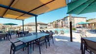 This expansive outdoor amenity space is designed to offer resort-style comfort and functionality within a modern residential community. The centerpiece is a large swimming pool surrounded by a generously sized travertine-tiled deck, lined with lounge chairs ideal for relaxing in the sun. Multiple shade structures with fabric canopies are strategically positioned over outdoor dining tables and chairs, providing relief from direct sunlight while maintaining an open, airy atmosphere under clear blue skies. A standout feature of the space is the built-in outdoor kitchen, constructed with a natural stone surround and equipped with a stainless steel grill, offering an ideal setup for outdoor cooking and entertaining. Neatly maintained landscaping with ornamental grasses and black mulched beds lines the perimeter, framed by black metal fencing that defines the space while preserving sightlines to the surrounding greenery. A paved walkway runs alongside the building, connecting various sections of the amenity area with ease. The multi-story residential buildings surrounding the space feature a warm mix of stucco, stone accents, and balconies, contributing to a cohesive and upscale community aesthetic. The overall ambiance is lively yet relaxed, purpose-built for outdoor gatherings and leisure.