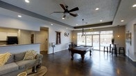 This single-floor community amenity space is thoughtfully designed to offer both recreation and relaxation. The main gathering area serves as the centerpiece, showcasing rich, polished concrete floors that lend an industrial yet refined aesthetic throughout. A full-size billiards table anchors the room, positioned beneath an elegant chandelier-style pendant light that adds a warm, social ambiance. A ceiling fan with dark wood-toned blades complements the recessed lighting, ensuring the space remains bright and well-ventilated. The textured ceiling adds subtle visual depth, while neutral gray walls provide a clean, modern backdrop. Large sliding glass doors span the far wall, flooding the interior with natural light and offering direct views and access to an outdoor pool area, seamlessly blending indoor and outdoor living. Decorative industrial-style wall accents, including gear motifs, contribute to the space's character. A seating area with comfortable furnishings offers a more relaxed retreat. An adjacent entryway with a contrasting door and a glimpse of a counter space suggests additional functional areas nearby. Overall, the space strikes a balance between casual comfort and active entertainment.