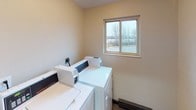 This single-floor space is purpose-built as a shared laundry facility, designed with practicality and functionality at its core. The room is finished with neutral, light-colored walls that contribute to a clean and utilitarian atmosphere throughout.

At the heart of the space are multiple Speed Queen commercial washers and dryers, arranged to maximize usability within the room. These heavy-duty machines are well-suited for high-frequency use, reflecting the space's intent as a shared or communal laundry area.

Natural light enters through sliding windows that overlook an exterior parking area with mature trees visible in the distance, lending a modest but welcome brightness to the room. Baseboard heating units run along the lower perimeter of the walls, ensuring the space remains comfortable during colder months.

The flooring consists of durable, low-maintenance carpeting in neutral tones. A utility access panel is mounted on one wall, providing convenient access to internal systems. A dark-framed interior door provides entry and exit, adding a subtle contrast to the light-toned walls.

Overall, the space is straightforward and efficiently organized, prioritizing ease of use and maintenance. It serves as a practical, well-equipped laundry room with the essential infrastructure to support regular commercial or communal laundry operations.