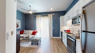 This thoughtfully designed single-floor residence showcases a cohesive interior palette anchored by striking navy blue accent walls, crisp white trim, and warm wood-tone flooring that flows seamlessly throughout the space. The open-concept layout connects the living area and kitchen, creating an inviting and functional environment. The kitchen is equipped with stainless steel appliances — including a range, over-the-range microwave, and dishwasher — complemented by white shaker-style cabinetry, granite countertops, under-cabinet lighting, and a decorative hexagonal tile backsplash. The living area offers a comfortable, relaxed atmosphere, highlighted by a ceiling fan, recessed lighting, and a sliding glass door with floor-to-ceiling vertical blinds that allow natural light to fill the room. The bedroom continues the navy blue accent theme, with warm lighting and a serene ambiance well-suited for rest. The bathroom features a granite-topped vanity with white cabinetry, a glass-enclosed shower, and direct access to a generously sized walk-in closet with built-in shelving and hanging rods. Every detail of this unit reflects a modern, polished aesthetic balanced with everyday practicality.