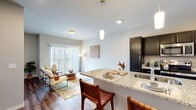 This single-floor residence offers a thoughtfully designed open-concept layout that seamlessly connects the kitchen, dining, and living areas. The kitchen is a standout feature, showcasing dark espresso cabinetry that contrasts beautifully with light granite countertops. A spacious peninsula with bar seating and pendant lighting serves as both a functional workspace and a casual dining hub. Stainless steel appliances, including a full-size refrigerator, range, and over-the-range microwave, complete the well-equipped cooking space. Rich wood-tone flooring flows throughout the main living area, adding warmth and cohesion to the open floor plan. The living area is bright and inviting, benefiting from natural light that streams in through sliding glass doors fitted with vertical blinds. The bedroom is a comfortable retreat, featuring plush carpeting, a ceiling fan, and a window that allows soft natural light to filter in, creating a calm and restful atmosphere. The bathroom is finished in a clean, contemporary style with a wide vanity, white countertop, large mirror, and a shower-tub combination, all complemented by neutral gray tones. Overall, this residence combines modern finishes with a functional layout, offering a comfortable and stylish living environment.