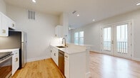 This well-appointed single-floor unit offers a thoughtfully designed open-concept layout that seamlessly integrates the kitchen and living area. Light hardwood flooring runs throughout the main living space, contributing to a warm and airy ambiance that is further enhanced by crisp white walls and recessed lighting.

The kitchen is a standout feature, equipped with white shaker-style cabinetry accented by brushed gold hardware, a white quartz countertop, a stainless steel undermount sink with a gold-tone faucet, and stainless steel appliances including a refrigerator and dishwasher. A peninsula counter extends outward, providing additional prep space and a casual dining or seating area that flows naturally into the living room.

The living area is generously proportioned relative to the overall footprint, benefiting from a ceiling fan, recessed lighting, and multiple windows that invite natural light. A glass-panel exterior door opens to an outdoor area, extending the sense of space and providing convenient access to the outside.

The bathroom is finished with elegant marble-look geometric tile flooring, a white vanity with a rectangular mirror, and clean, neutral walls that maintain the home's cohesive modern aesthetic. A dedicated closet provides practical storage. The overall design is clean, contemporary, and move-in ready.