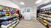Located at 186 E Future Way, this compact convenience store is thoughtfully designed to deliver a full retail experience within an efficient single-level footprint. The space features a clean, modern aesthetic with crisp white walls, a drop-ceiling with recessed panel lighting, and durable patterned vinyl plank flooring in neutral tones of gray and gold throughout.

Along one wall, a row of large glass-door commercial refrigerators showcases a well-stocked selection of beverages and frozen food items. Complementing these, a dedicated chest freezer unit prominently branded for ice cream provides additional frozen treat options. Dark, freestanding display shelving units line the walls and central areas, organizing a diverse range of snacks, packaged goods, and everyday essentials.

A self-checkout kiosk is conveniently positioned alongside the shelving, facilitating quick and easy transactions. A wall-mounted monitor displays promotional or instructional content to assist shoppers. The store benefits from two access points: a glass sliding door that connects directly to an adjacent parking garage, and a secondary door providing an alternate entry and exit route.

Framed artwork decorates the corridor walls, adding a polished touch to the otherwise functional space. Security cameras are discreetly integrated throughout, ensuring a safe shopping environment. The overall layout prioritizes ease of navigation and accessibility, making this a well-appointed and practical retail destination.