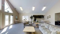 This single-floor space is designed as a multifunctional gathering area, characterized by a prominent vaulted ceiling that enhances the sense of openness and airiness throughout. Flush-mounted fluorescent light fixtures run across the ceiling, providing consistent, even illumination throughout the room.

The layout is divided into distinct functional zones that flow seamlessly into one another. The central lounge area is furnished with floral-patterned upholstered sofas and armchairs arranged around low wooden coffee and side tables, creating a comfortable seating arrangement suited for socializing. A decorative potted tree adds a natural element to the space.

Along the perimeter, dining areas feature lightweight chairs and tables positioned near a window that allows natural light to filter in. The background reveals a dark-countered kitchenette equipped with a refrigerator and cabinetry, suggesting the space supports catered gatherings or self-serve refreshments.

The flooring is covered in neutral gray carpet throughout the main areas, complementing the beige and white color palette of the walls and furnishings. The overall ambiance is clean, functional, and welcoming, well-suited for use as a community or common room.