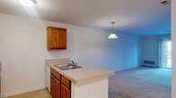 This single-floor residence offers a functional and well-organized layout designed for comfortable everyday living. The kitchen serves as a central feature, equipped with warm oak cabinetry, light laminate countertops, a double stainless steel sink set into a peninsula, and white appliances including a refrigerator, electric range, and over-the-range microwave. The peninsula provides a natural division between the kitchen and the adjoining living area, which is finished with neutral carpet and benefits from a sliding glass door that invites natural light and provides outdoor access. An additional bedroom features a large multi-pane window with views of the surrounding exterior, further enhancing the sense of openness throughout. The full bathroom is cleanly finished with large-format tile flooring, an oak vanity with a single sink, a wide wall mirror with overhead lighting, and a standard tub-shower combination. A dedicated laundry room houses a full-sized washer and dryer side by side, complete with wire shelving above — a practical and convenient amenity. Tile flooring in the kitchen and utility areas contrasts with soft carpeting in the living and sleeping spaces, creating a cohesive and livable environment throughout.