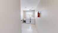Located at 840 Trillium Pk in Sarnia, Ontario, this single-floor laundry room is a compact, functional utility space designed to serve its users efficiently. The room features light-coloured walls and speckled vinyl tile flooring that contribute to a clean, practical aesthetic throughout.

The laundry area is equipped with a top-loading washing machine and a full-sized dryer positioned side by side, along with a utility sink for added convenience. A window above the machines allows natural light to filter in, creating a brighter and more pleasant environment. A wall-mounted folding shelf provides a dedicated surface for sorting and folding laundry, making the most of the available space.

A notice board displaying safety information and laundry rules is mounted on the wall, keeping users informed of guidelines and operating hours. Additional practical features include a fire extinguisher near the entry door and a small waste bin. The entry corridor leads to a solid door with a small window, offering secure and straightforward access to the room. Intercom and mailbox panels are also visible near the entrance, suggesting this laundry room is part of a larger residential building. Overall, the space is thoughtfully organized to maximize utility within its modest footprint.
